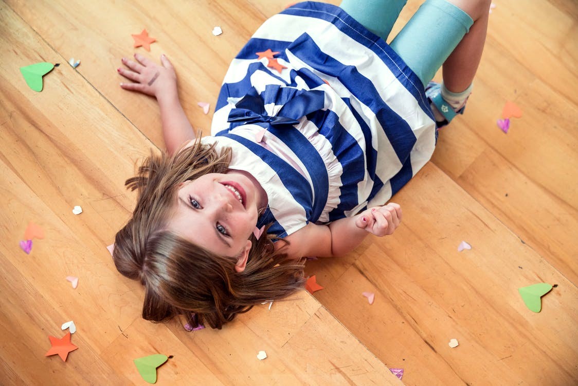 A little girl lying on the wooden floor with confetti spread around her.