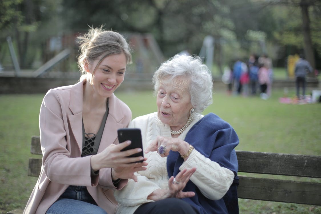 A young woman sitting on a bench with an old lady and showing something on the phone.
